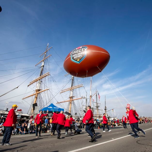 Men in red suits dance while holding strings to a large football balloon emblazoned with “DIRECTV Holiday Bowl San Diego.”