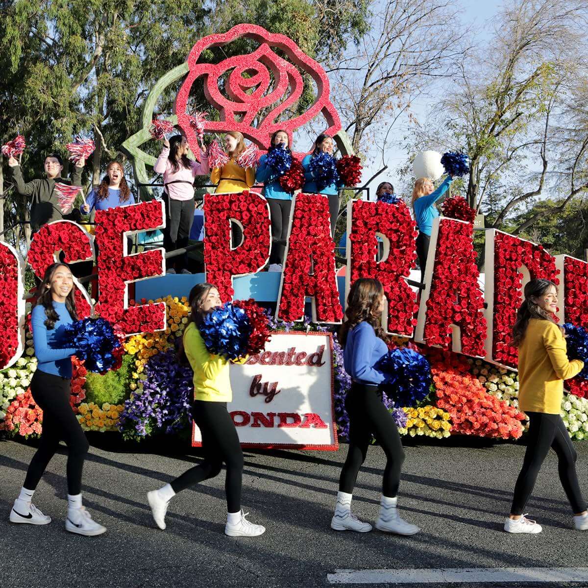 A group of cheerleaders walk with the official “Rose Parade” float, decorated with red roses and other colorful flowers.