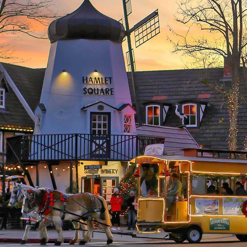 Hamlet Square in Solvang, illuminated by holiday lights. A festive horse-drawn bus is making its way across the road.