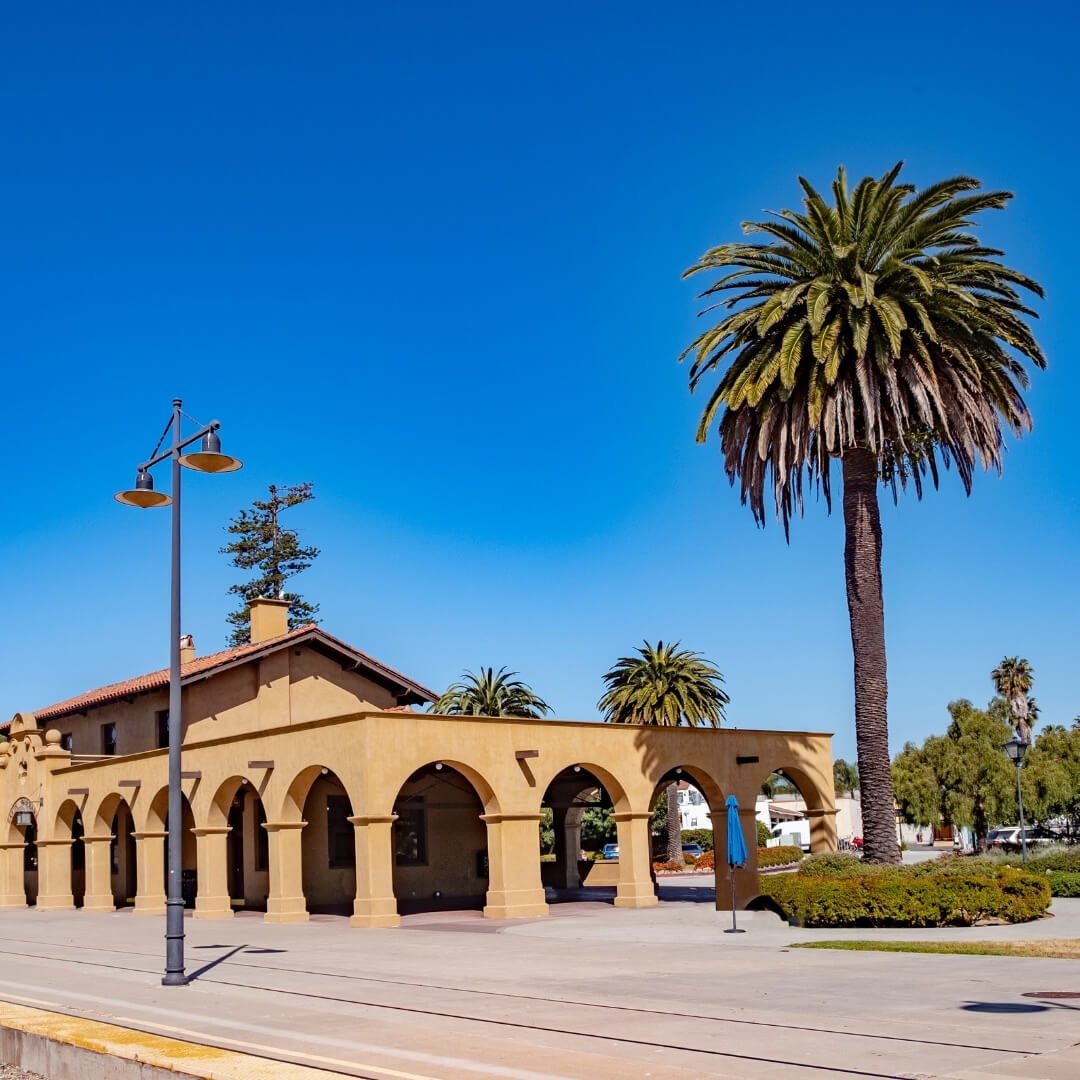 A view from the side of the Santa Barbara Amtrak Station, slightly shaded by a large palm tree.